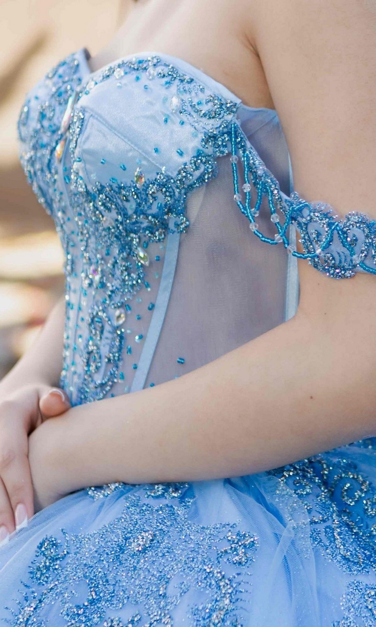 Close-up of a person wearing the Quinceanera Dress 8190J By Cinderella Couture, a blue beaded strapless gown featuring sheer fabric and intricate glittery embellishments, evoking an enchanting off-the-shoulder ball gown style.